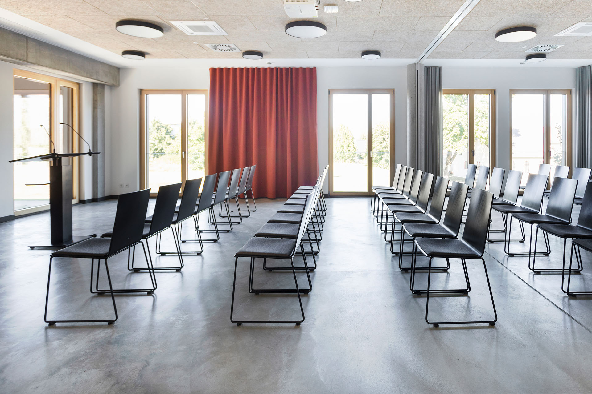 Konferenzraum Seitenansicht Konferenzraum zum Mieten mit Reihenbestuhlung und Rednerpult vor großen Fenstern und rotem Vorhang