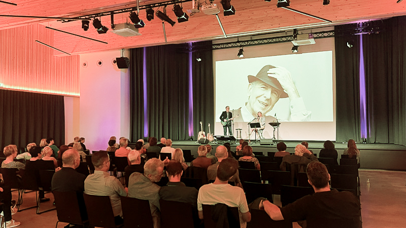 Saal Konzertbeispiel Saal zum Mieten mit Musiker auf großer Bühne und vielen Menschen auf schwarzen Stühlen