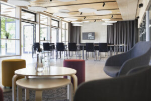 Foyer „Mietek“ helles Foyer mit Sesseln und kleinen Tischen im Vordergrund und Stühlen, großen Tischen, Bildschirm und Vorhängen im Hintergrund
